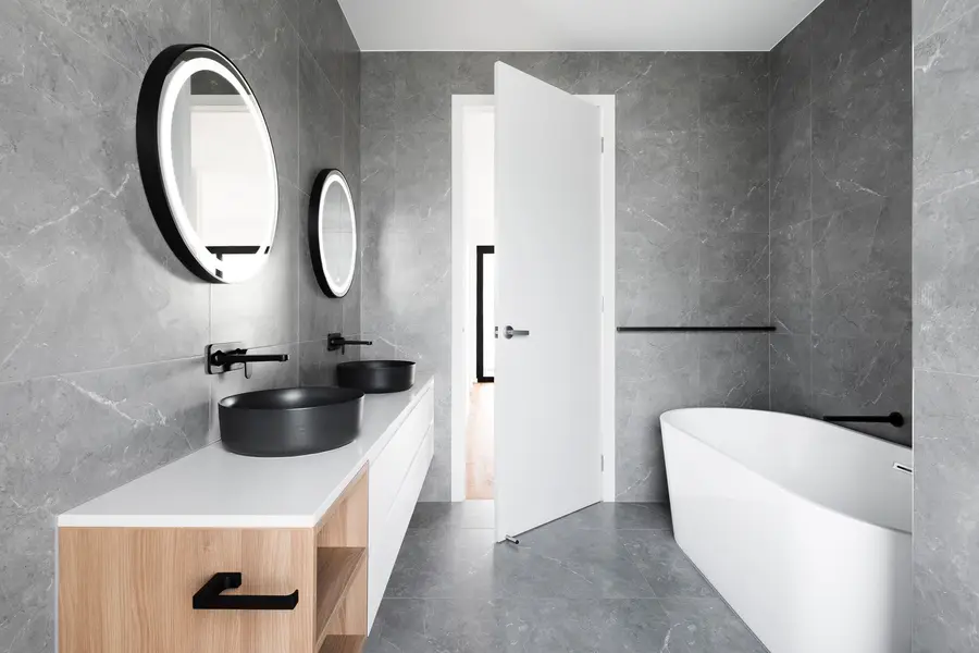 Modernes Badezimmer mit doppelten schwarzen Waschbecken, ovalen beleuchteten Spiegeln, weißer Badewanne und grauen Fliesen.