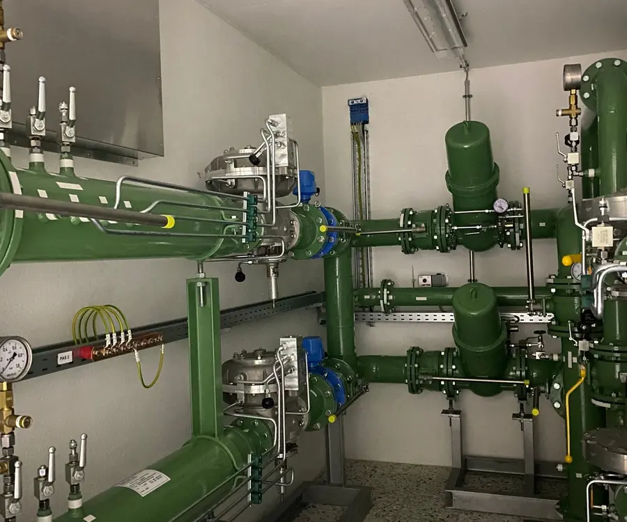 Raum mit komplexer Anordnung grüner Rohre, Ventile und Messgeräte an weißen Wänden und Decke.
