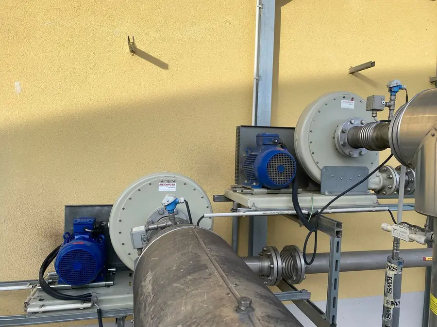 Zwei Industriegebläse mit blauen Elektromotoren an einer gelben Wand montiert, verbunden mit Metallrohren.
