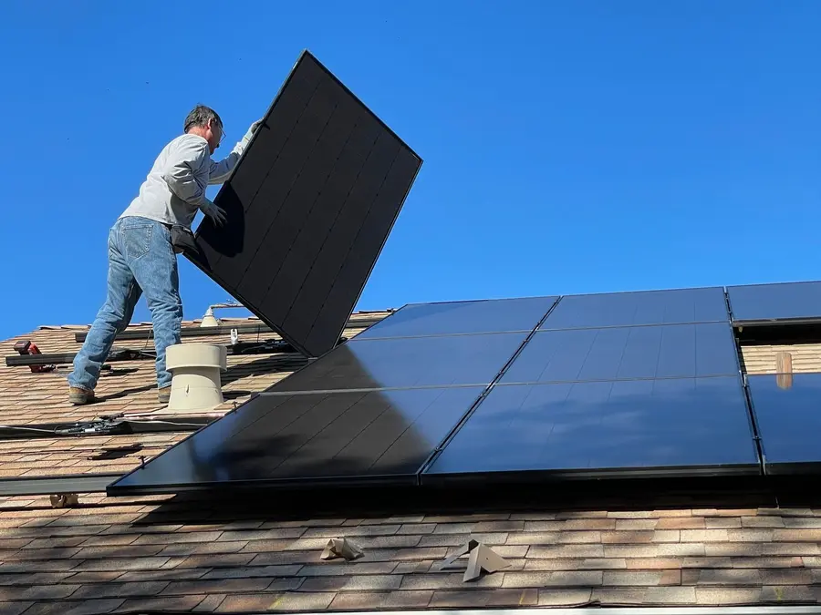 Ein Mann installiert ein schwarzes Solarmodul auf einem braunen Ziegeldach unter blauem Himmel.